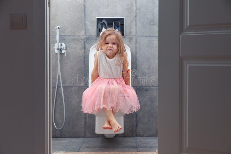 Cute little girl in white pink dress sitting on toilet with toilet paper on background of walls with gray tiles, view. From open door stock photos