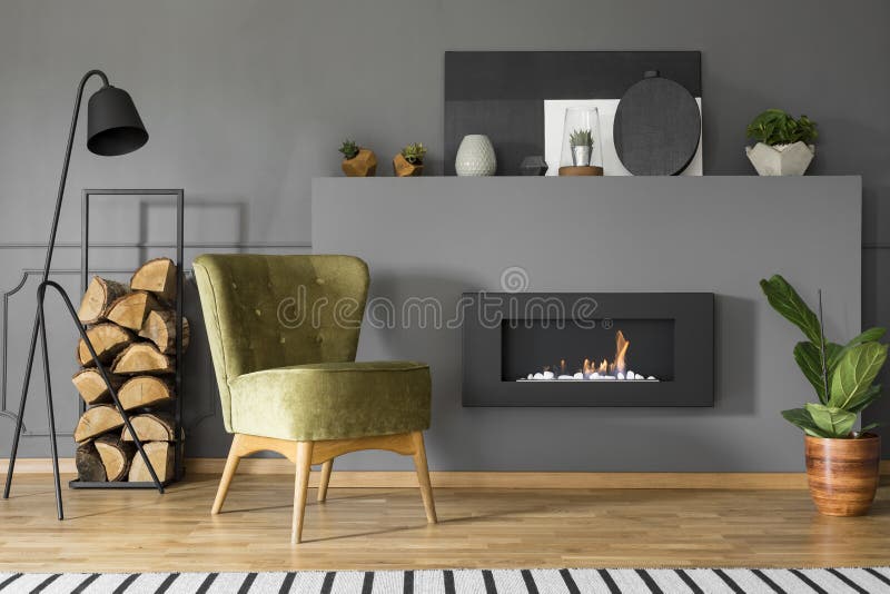 Green armchair and firewood next to fireplace in grey living room interior with lamp. Real photo stock photography