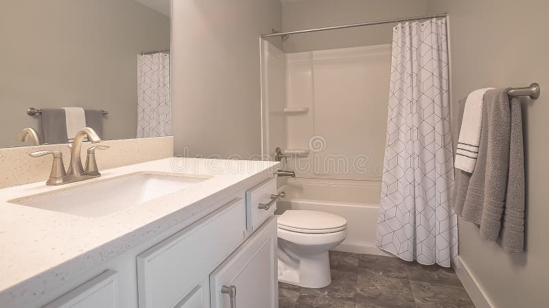 Panorama Home bathroom interior with dark gray tile floor and light gray wall. The room is furnished with a vanity unit, toilet, and bathtub with shower and stock image
